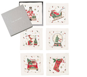 The Kim Seybert Holly Jolly Cocktail Napkin Set includes six square white linen napkins, each with a unique embroidered Christmas motif, shown beside a gray Kim Seybert box.