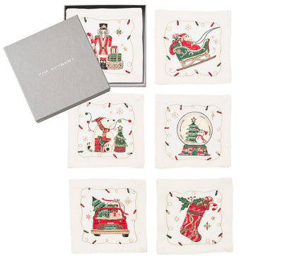 The Kim Seybert Holly Jolly Cocktail Napkin Set includes six square white linen napkins, each with a unique embroidered Christmas motif, shown beside a gray Kim Seybert box.