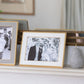 A black-and-white wedding photo in the Addison Ross Wide Curved Enamel Frame Collection is displayed on a mantel beside another framed picture and a decorative stone object, featuring a soft velvet backing for an elegant touch.