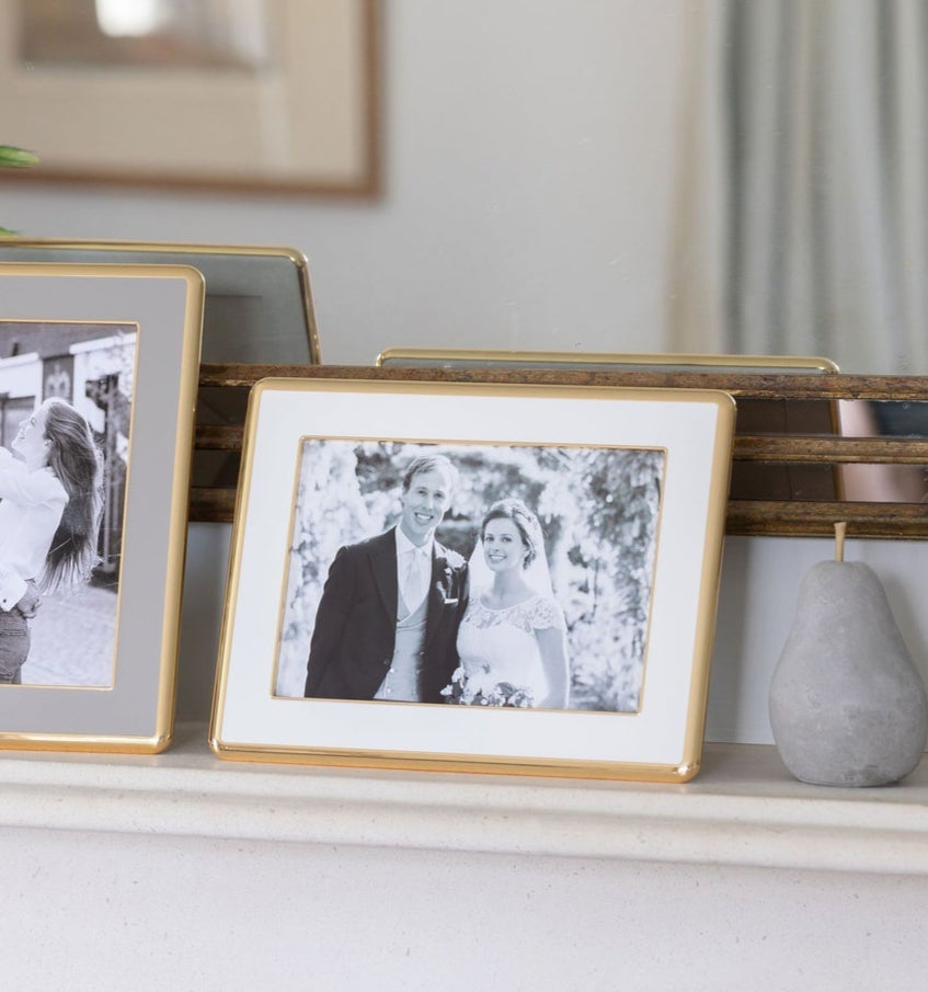 A black-and-white wedding photo in the Addison Ross Wide Curved Enamel Frame Collection is displayed on a mantel beside another framed picture and a decorative stone object, featuring a soft velvet backing for an elegant touch.