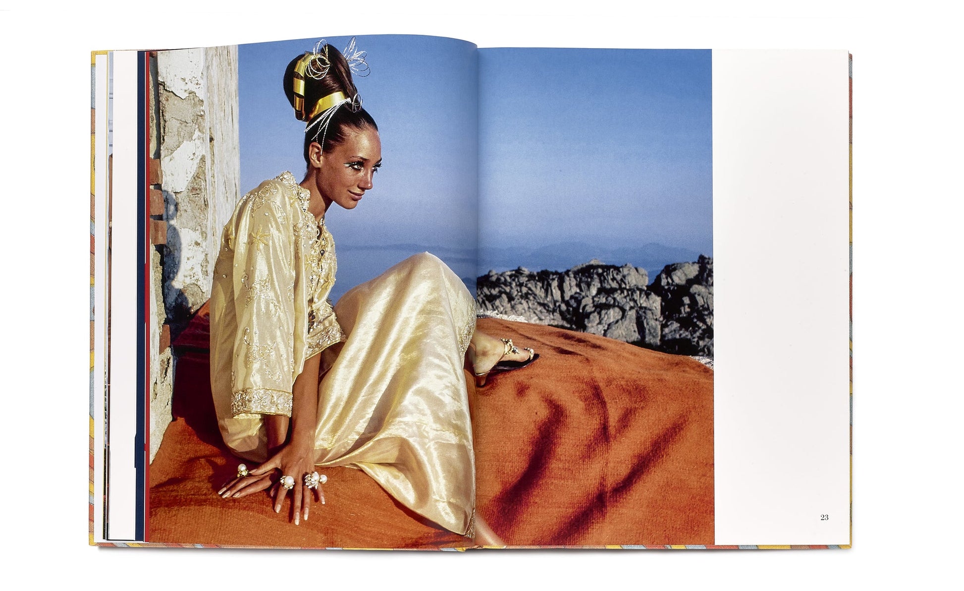 Draped in Vendome's Caftans: From Classical to Camp and a lavish headpiece, a person sits elegantly on an orange blanket against the breathtaking mountainous backdrop—a perfect resort setting for this display of style.
