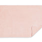 A light pink rectangular bath mat from the Matouk Cairo Scallop Bath Collection, featuring scalloped edges and a white trim reminiscent of luxury towels; one corner is partially flipped to reveal the underside.
