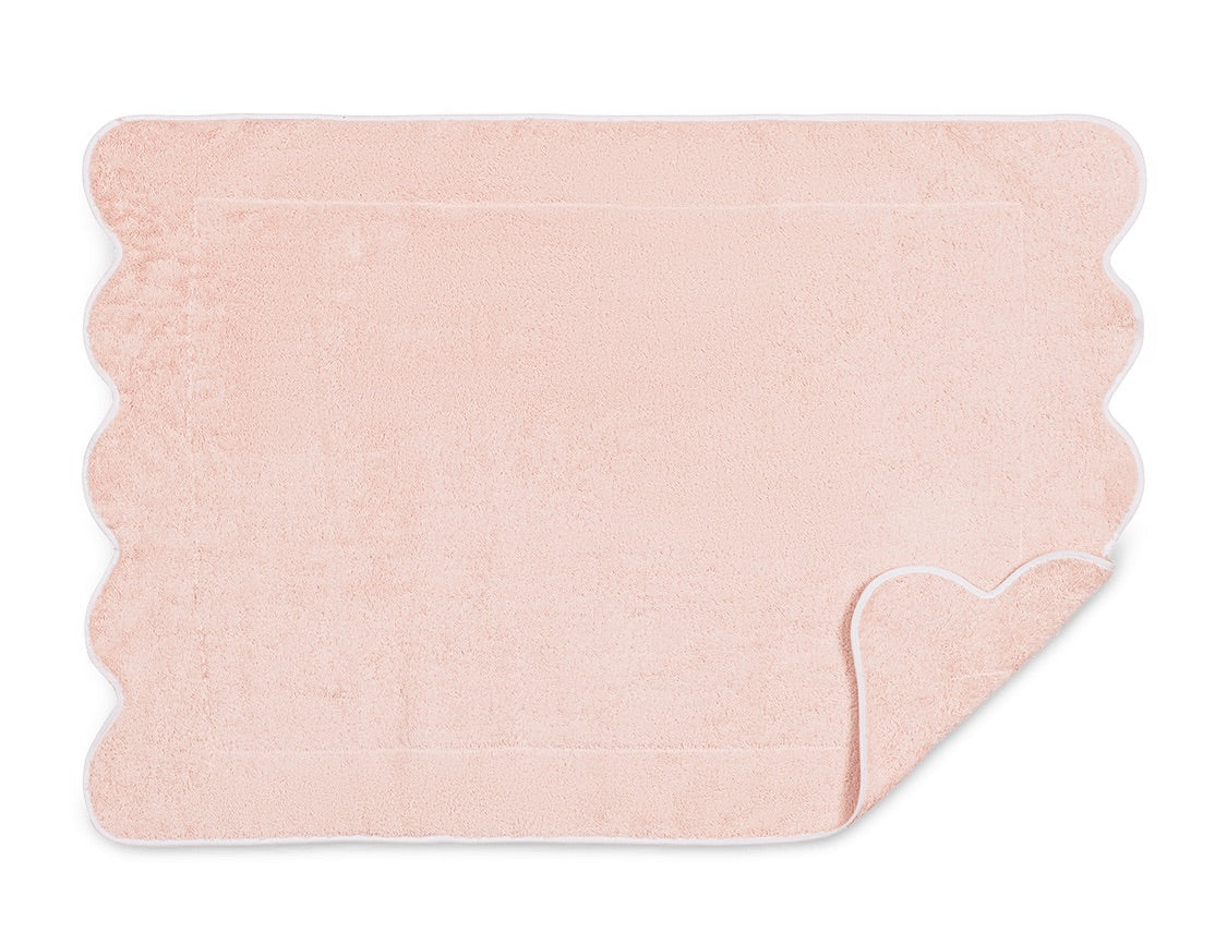 A light pink rectangular bath mat from the Matouk Cairo Scallop Bath Collection, featuring scalloped edges and a white trim reminiscent of luxury towels; one corner is partially flipped to reveal the underside.
