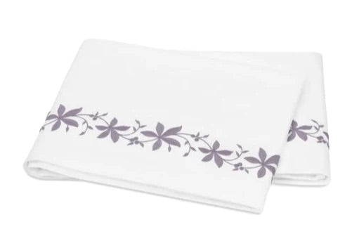 A folded white bed sheet from the Matouk Callista Bedding Collection, expertly crafted from luxurious Giza Egyptian cotton with a 500 thread count, showcasing a delicate purple floral pattern and decorative embroidery along one edge, displayed on a plain background.