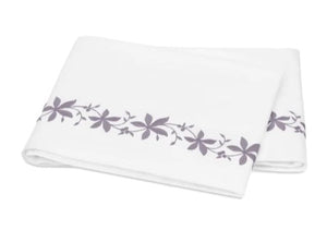A folded white bed sheet from the Matouk Callista Bedding Collection, expertly crafted from luxurious Giza Egyptian cotton with a 500 thread count, showcasing a delicate purple floral pattern and decorative embroidery along one edge, displayed on a plain background.