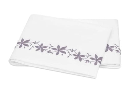A folded white bed sheet from the Matouk Callista Bedding Collection, expertly crafted from luxurious Giza Egyptian cotton with a 500 thread count, showcasing a delicate purple floral pattern and decorative embroidery along one edge, displayed on a plain background.