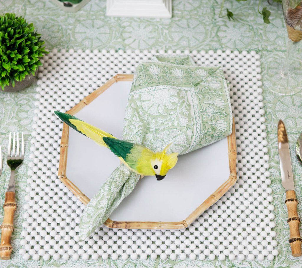 A place setting with a hexagonal plate, Kim Seybert Provence Napkin Set, parrot-shaped napkin ring, beaded placemat, bamboo-handled flatware, matching tablecloth, and a green plant nearby.