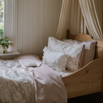 A wooden bed dressed in the Gooselings Bows & Butterfly Kisses Toddler Duvet sits by a window with cream curtains and pastel bedding, complete with pillows and a flower-filled vase nearby.
