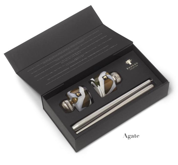 An open black box showcases two intricately designed hand-blown glass bottle stoppers, accompanied by a metal rod labeled "Agate," reflecting the elegance of the Baobab Collection Candle Holder Twins by Baobab.