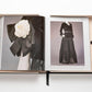 Open book showcasing fashion photographs. Left page features a close-up of a black outfit with a white flower accessory; right page shows a full-length black couture outfit with text above it, channeling luxe minimalism. The Impossible Collection of Chanel by Assouline placed in a light-colored box.