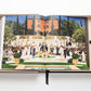 An open book displaying a colorful fashion runway show in front of a grand building with columns and a staircase, surrounded by palm trees and garden foliage, embodies understated luxury reminiscent of Gabrielle Chanel's timeless elegance. The book is titled "The Impossible Collection of Chanel" by Assouline.