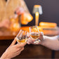 Two people clink Simon Pearce Windham Tasting Glass Gift Set glasses at a bar, with a bartender and a small lamp in the background.