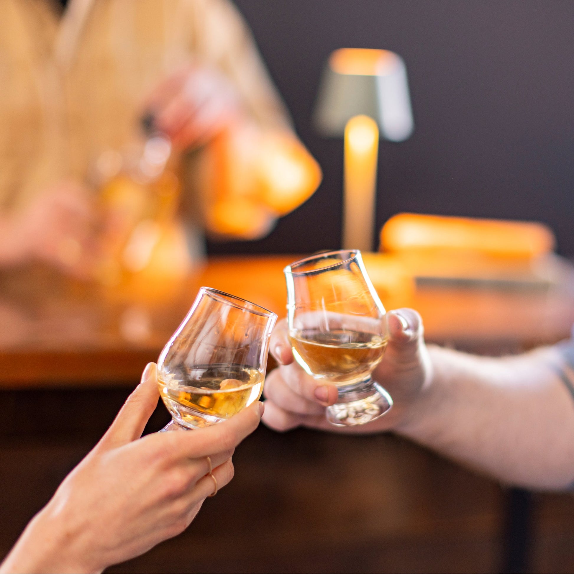 Two people clink Simon Pearce Windham Tasting Glass Gift Set glasses at a bar, with a bartender and a small lamp in the background.