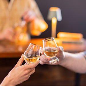 Two people clink Simon Pearce Windham Tasting Glass Gift Set glasses at a bar, with a bartender and a small lamp in the background.