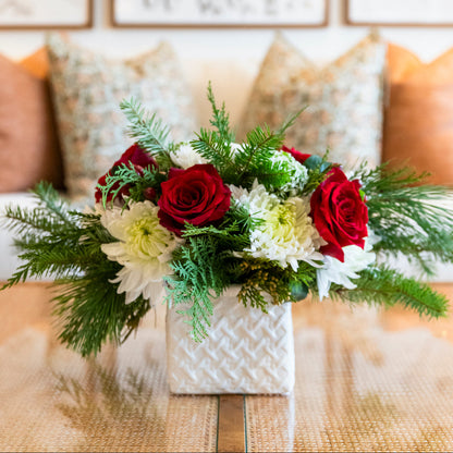 The Live Evergreen Holiday Floral Arrangement by Hive Floral Studio features festive red roses and seasonal blooms in a square white vase, displayed on a glass-topped table with pillows in the background.