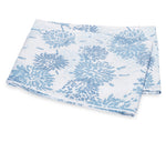 The Tillett x Matouk Chrysanthemum Bedding Collection in blue by Matouk showcases folded white fabric with a blue chrysanthemum floral motif, displayed on a plain background.