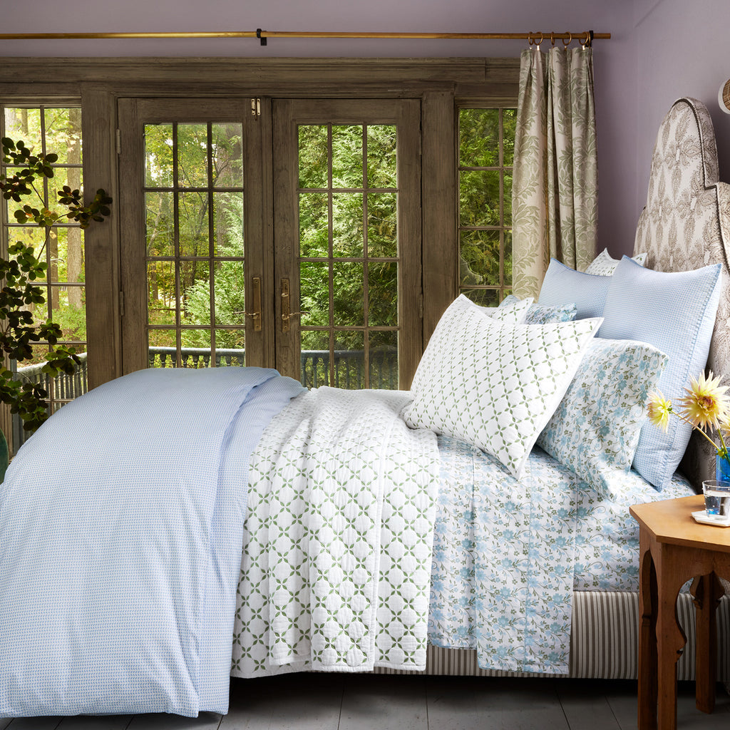 A neatly made bed with the John Robshaw Mihana Mist Bedding Collection in blue and white organic cotton sits near glass doors overlooking a green garden. A wooden nightstand with flowers beside the bed adds botanical harmony.