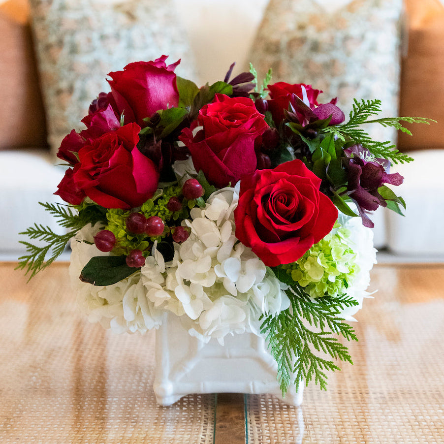 The Live Classic Holiday Floral Arrangement by Hive Floral Studio features red roses, white hydrangeas, green foliage, and berries in a square ceramic cachepot, making a festive centerpiece for your table.