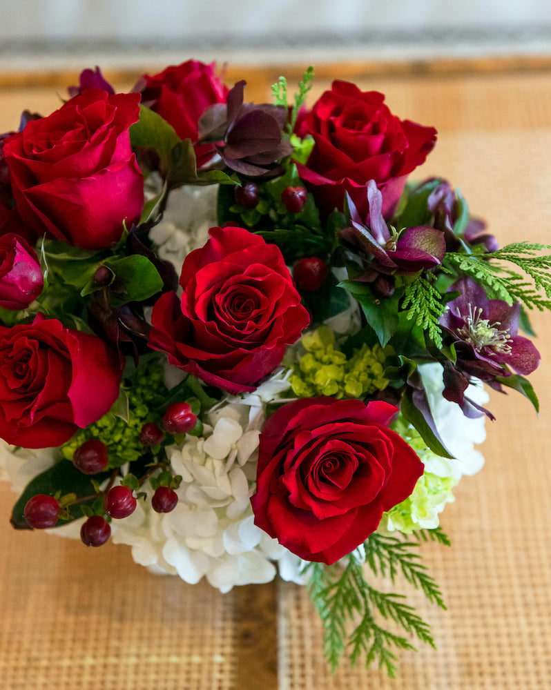 The Hive Floral Studio Live Classic Holiday Floral Arrangement is a festive centerpiece with red roses, white hydrangeas, dark berries, and lush greenery in a round ceramic cachepot on a woven surface.