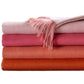 Four Classic Herringbone Throw blankets by Alicia Adams Alpaca, in pink and orange hues, are stacked with the top featuring fringed edges—each fair-trade throw is made in Peru for cozy, ethical comfort.