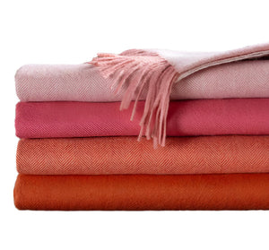 Four Classic Herringbone Throw blankets by Alicia Adams Alpaca, in pink and orange hues, are stacked with the top featuring fringed edges—each fair-trade throw is made in Peru for cozy, ethical comfort.