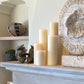 Five cream-colored candles from the Perin Mowen Connoisseur Pillar Candle Collection are arranged on a mantel, forming a natural centerpiece beside sculptural decor and a patterned vase displayed on the shelf in the background.