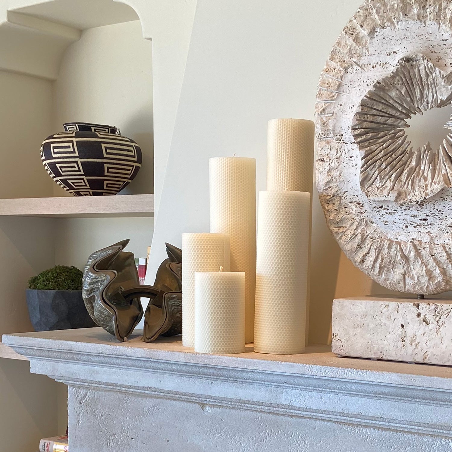 Five cream-colored candles from the Perin Mowen Connoisseur Pillar Candle Collection are arranged on a mantel, forming a natural centerpiece beside sculptural decor and a patterned vase displayed on the shelf in the background.