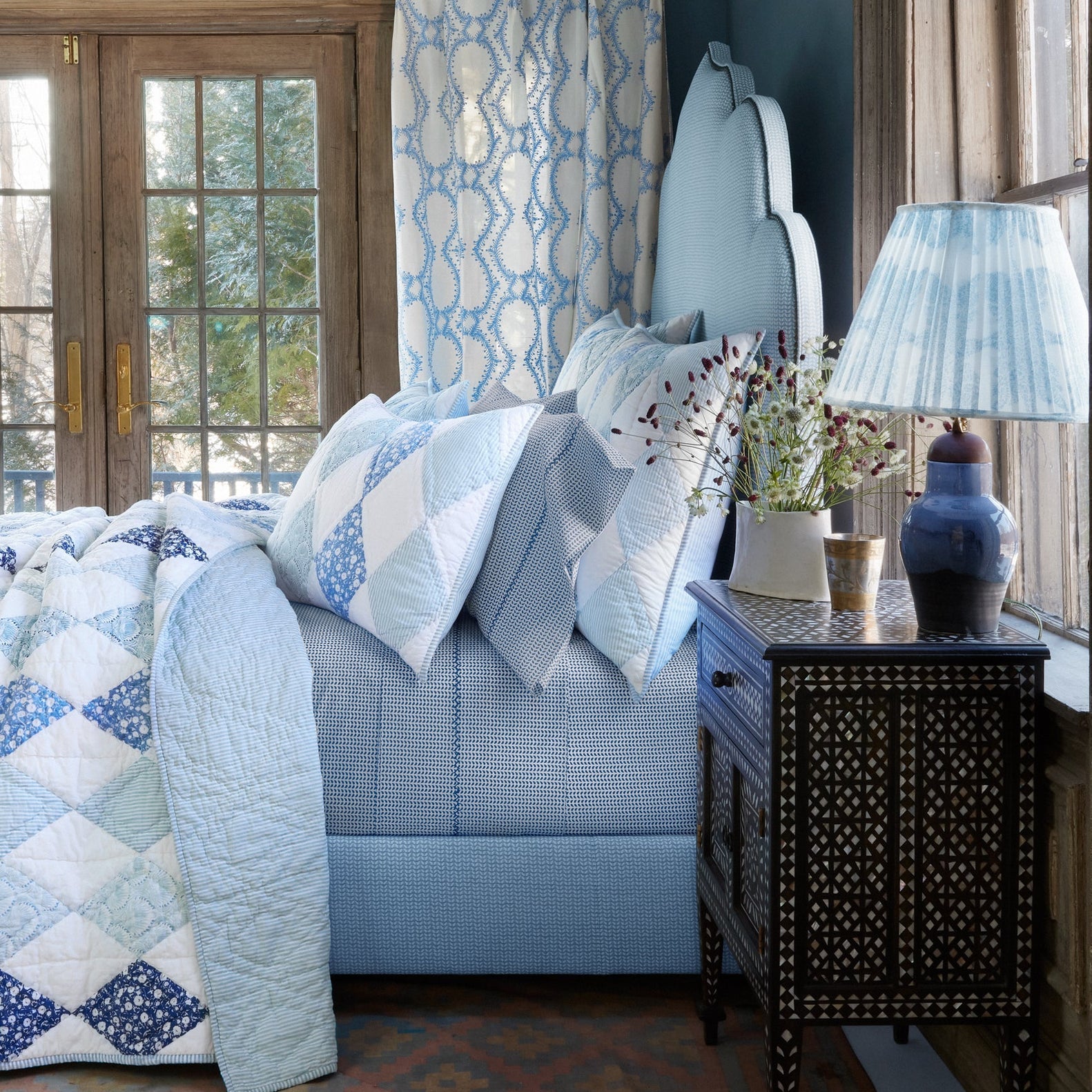 A blue and white bedroom featuring the John Robshaw Cinde Bedding Collection, including an organic sheet set in Light Indigo, an upholstered headboard, a side table with a lamp, and large windows that fill the space with natural light.