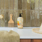 A bathroom countertop with a towel, wooden comb, liquid soap dispenser, and straw hat sits by glossy greenish-brown tiles and a gold faucet—evoking the fresh scent of Baobab’s Saint Tropez Body Wellness Collection.