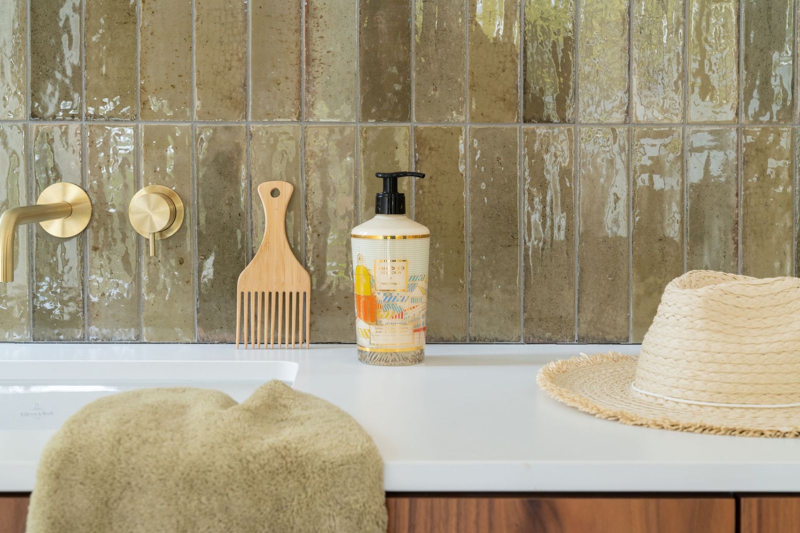A bathroom countertop with a towel, wooden comb, liquid soap dispenser, and straw hat sits by glossy greenish-brown tiles and a gold faucet—evoking the fresh scent of Baobab’s Saint Tropez Body Wellness Collection.