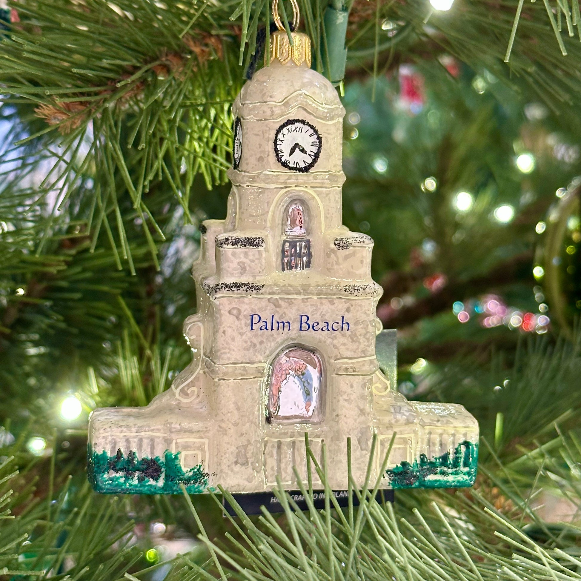 The Palm Beach Clock Tower Custom Ornament by Joy To The World Collectibles, Inc. hangs on a Christmas tree decorated with green needles and lights.