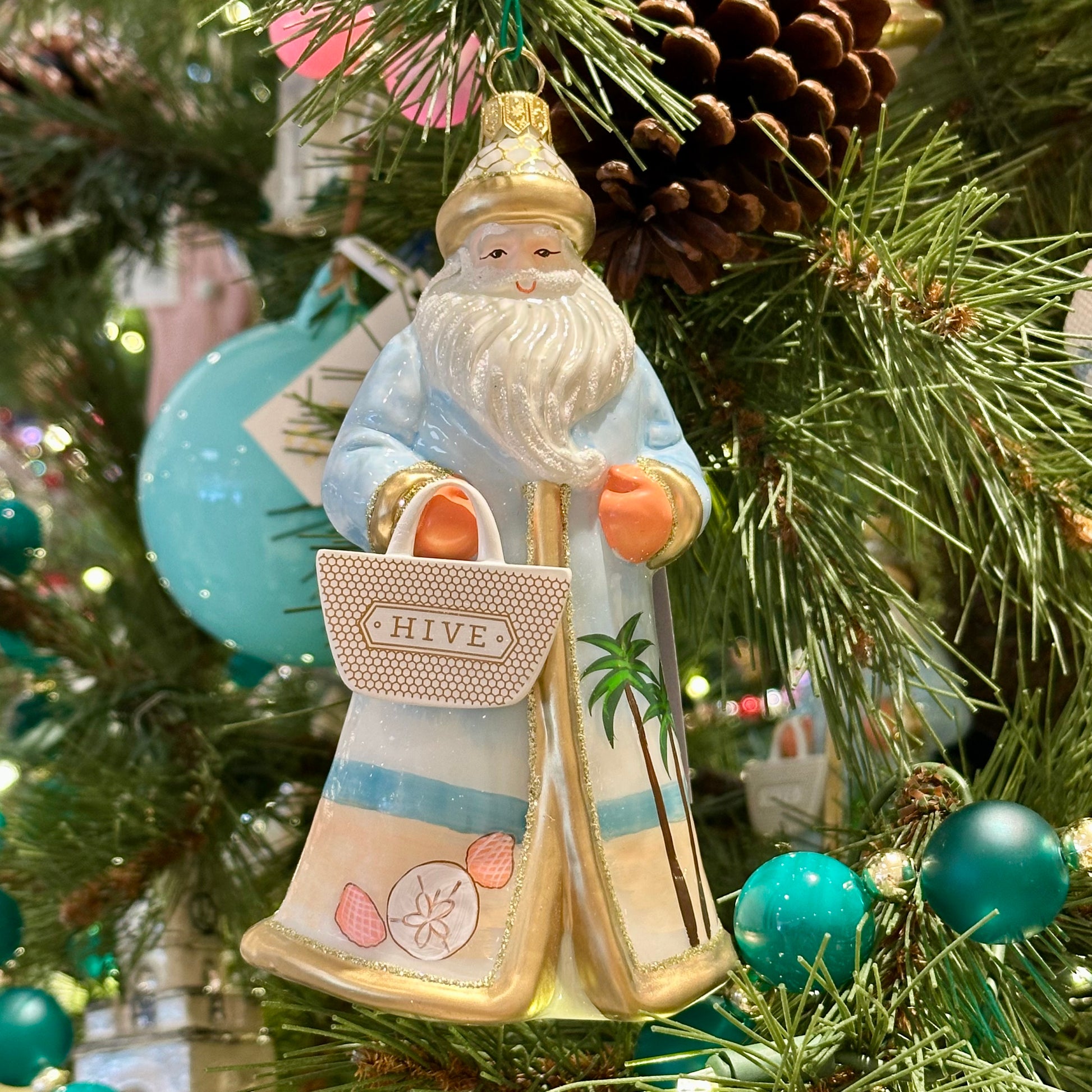 The Hive Santa Custom Ornament by Joy To The World Collectibles, Inc. features a sun-soaked Santa in blue and white with beachy details, holding a "HIVE" bag, hanging on a festive tree with pinecones and teal ornaments.