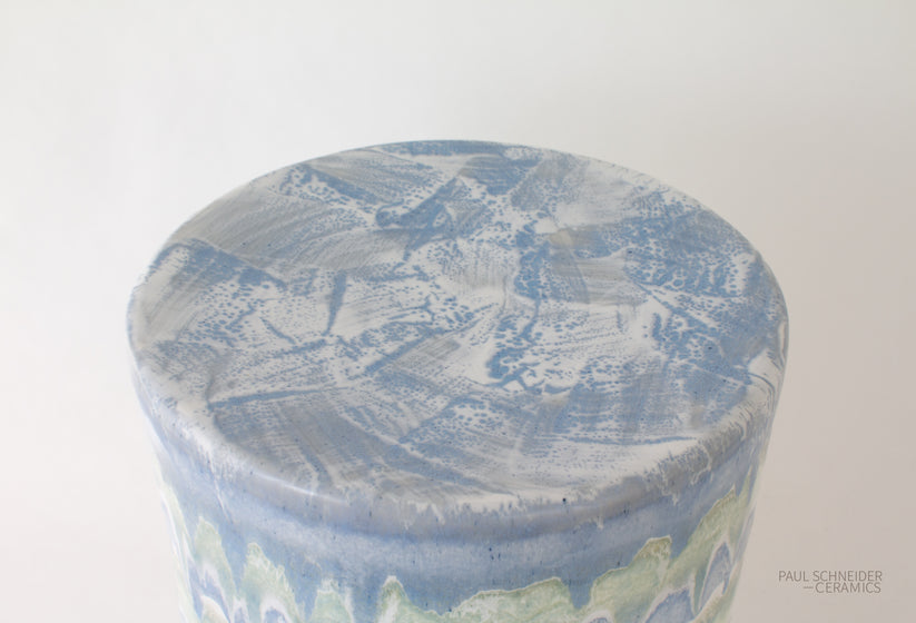 The Paul Schneider Ceramics Cylindrical Garden Stool Collection features a round ceramic piece with a blue and white marbled surface, subtle wavy green pattern on the side, and showcases unique variations, set against a white background.