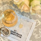 A glass of white wine and an oyster sit atop August Morgan's "Light Snack Heavy Pour" cotton cocktail napkin, with a bouquet of white roses in the background.