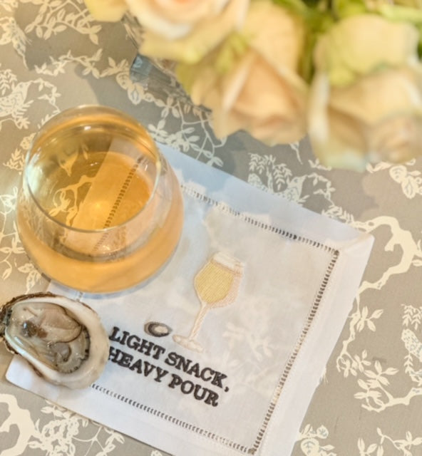 A glass of white wine and an oyster sit atop August Morgan's "Light Snack Heavy Pour" cotton cocktail napkin, with a bouquet of white roses in the background.
