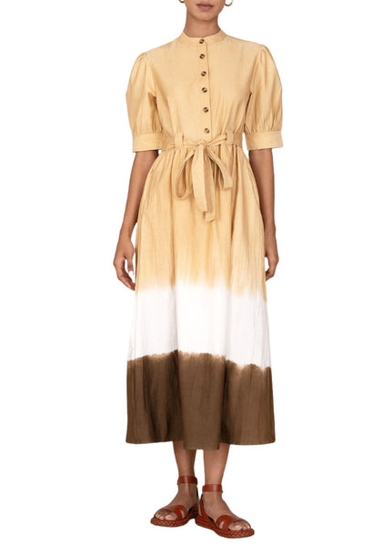 A person wearing the Banjanan Daffodil Dress with short puff sleeves, a buttoned front, and a waist tie belt. The cotton sateen fabric gives it an elegant sheen. They are also wearing brown sandals.