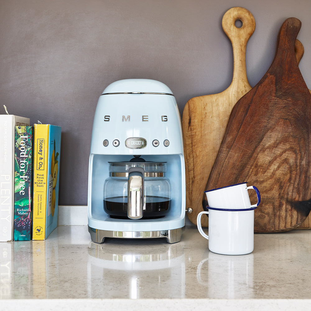 A SMEG Drip Filter Coffee Machine Collection in blue graces the kitchen counter beside a white mug. Wooden cutting boards and colorful cookbooks provide a backdrop, while a reusable coffee filter offers convenience and sustainability.