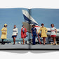 Six women in colorful uniforms pose before a Delta Air Lines plane on the tarmac, featured in "Delta: 100 Years and Climbing" by Assouline, an open airline history book celebrating Delta's centennial.