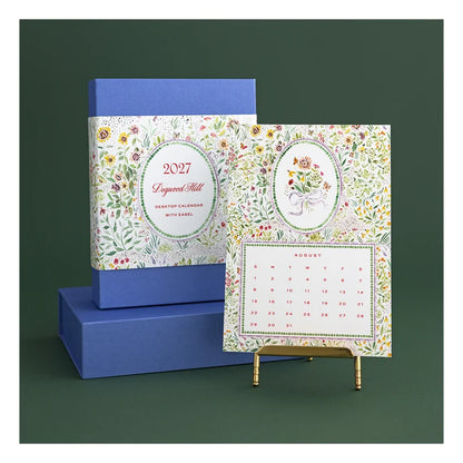 A Dogwood-Hill Desktop Calendar with Easel featuring a floral border, displayed on a gold wire easel next to its blue presentation box.
