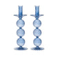 The Iris Tall Candle Holder Set by Kim Seybert features two identical blue, hand-blown glass holders with ribbed, spherical, and fluted designs and elegant scalloped bases, displayed side by side on a white background.