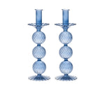 The Iris Tall Candle Holder Set by Kim Seybert features two identical blue, hand-blown glass holders with ribbed, spherical, and fluted designs and elegant scalloped bases, displayed side by side on a white background.