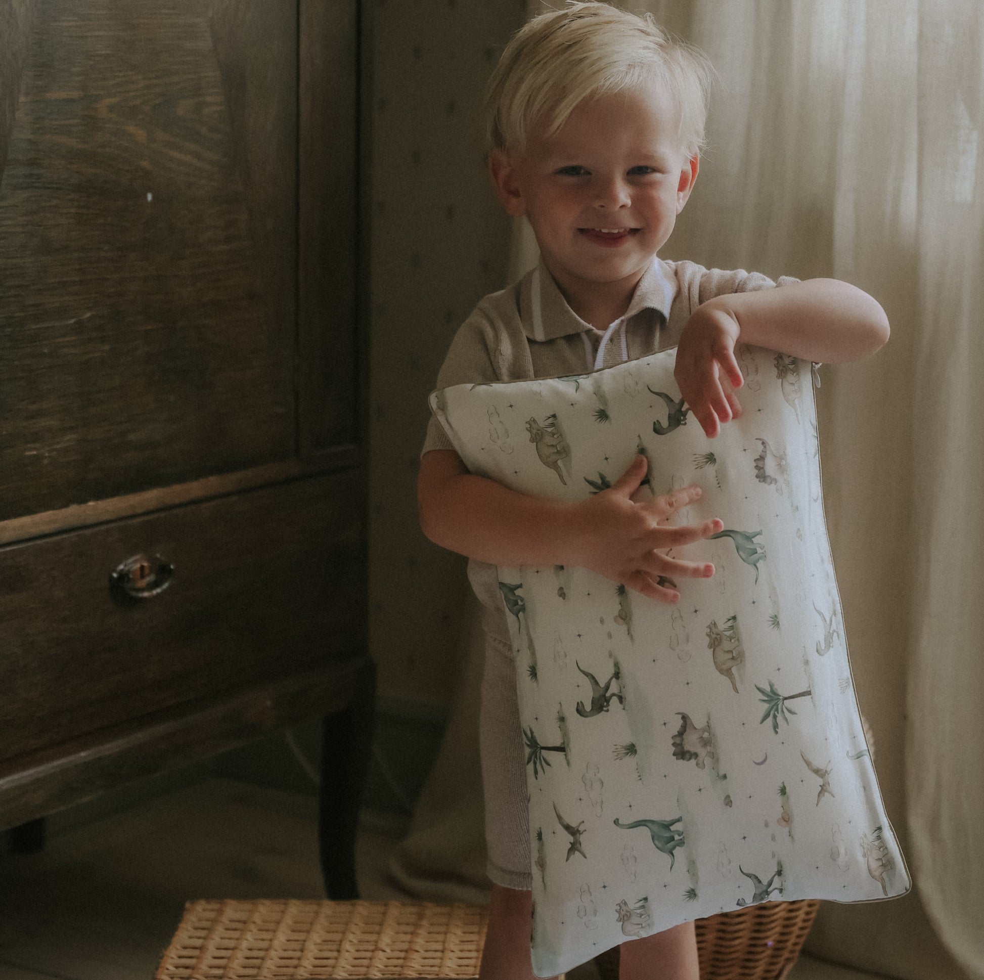 A young child with blonde hair smiles indoors, holding the Gooselings Dino Dreams Toddler Pillow with animal prints—perfect for sweet dreams.