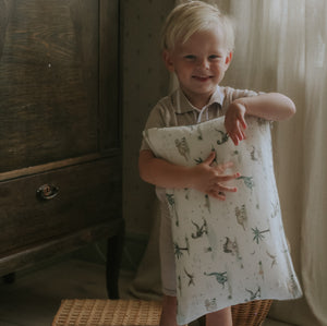 A young child with blonde hair smiles indoors, holding the Gooselings Dino Dreams Toddler Pillow with animal prints—perfect for sweet dreams.