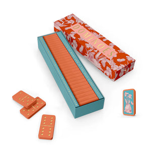 The Good Juju Ink Domino Set features orange dominoes with yellow dots in a decorative keepsake box, partially open to display neatly stacked tiles—three dominoes and the lid are displayed beside it, making it a perfect gift for design lovers.