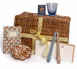 The Chic Gift Hamper by Sharland England features a wicker basket with yellow ribbon, patterned notepad, ceramic dish, sustainable soap, hand-poured candles, blue glass, and notecards—ideal for artisanal gift lovers.