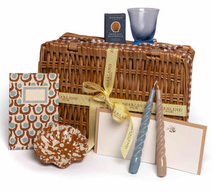 The Chic Gift Hamper by Sharland England features a wicker basket with yellow ribbon, patterned notepad, ceramic dish, sustainable soap, hand-poured candles, blue glass, and notecards—ideal for artisanal gift lovers.