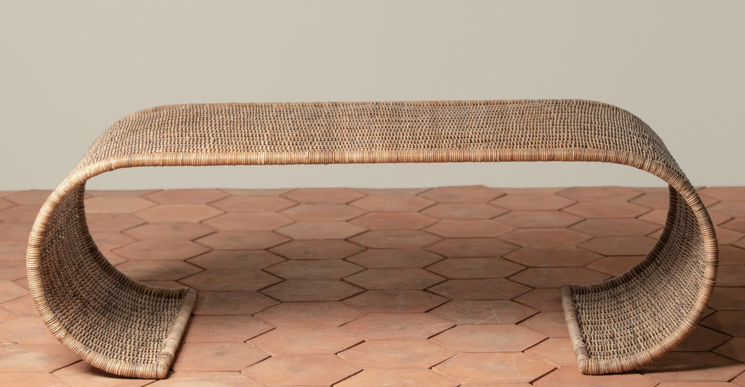 The Selamat Healdsburg Wicker Coffee Table, crafted from sustainably harvested rattan with curved ends, sits on a terracotta hexagon tile floor against a plain light-colored wall.