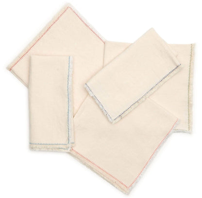 The Nomad Stitch Fringe Napkin Set by Deborah Rhodes features five off-white napkins with frayed edges and vibrant blue or red stitching along one side, arranged overlapping on a plain background.