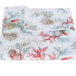 The Matouk Del Tesoro Bedding Collection in Coral features a white bedsheet decorated with vibrant red, blue, green, and beige coral and seashells, neatly folded with one corner artfully turned over.