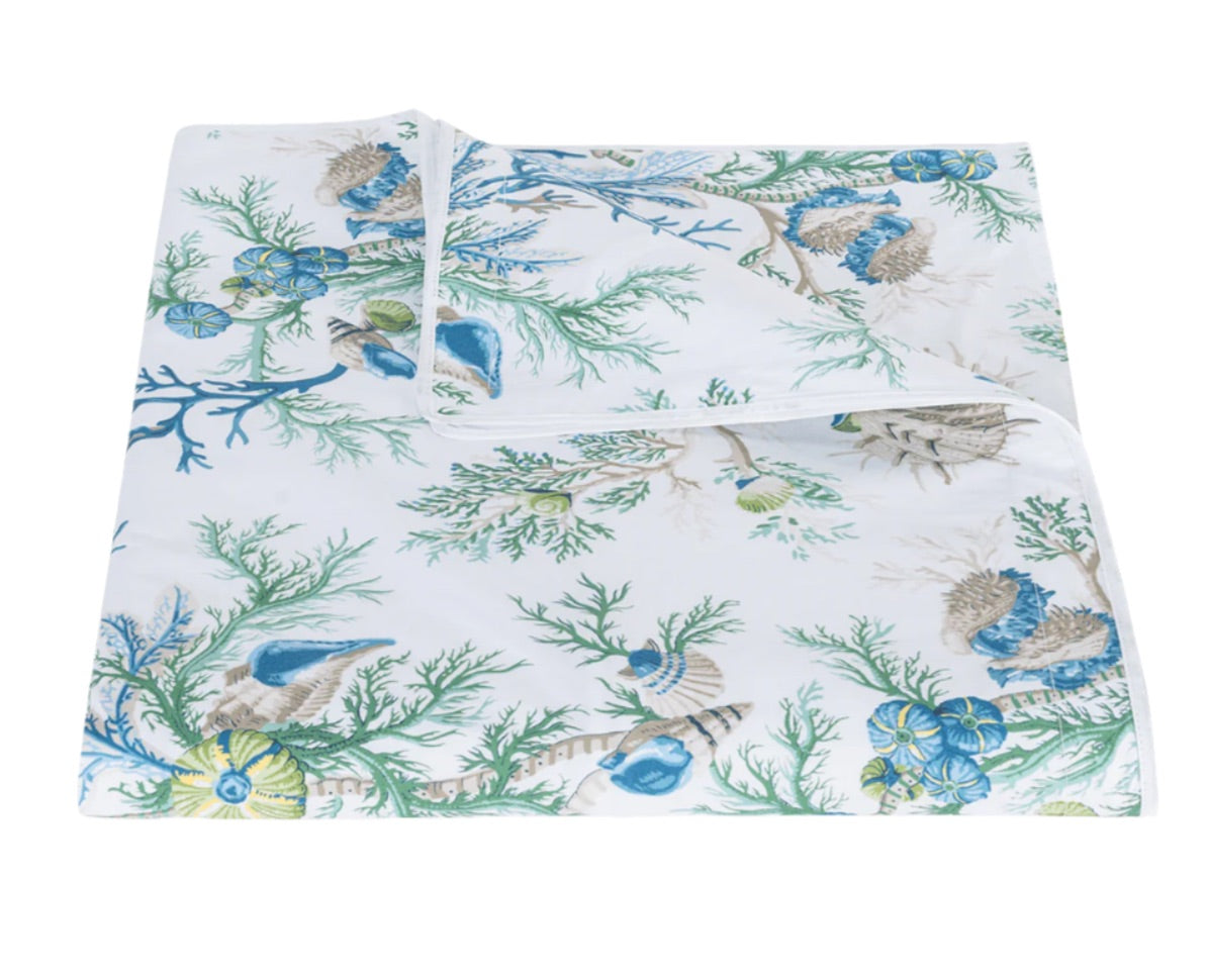 Partially unfolded to reveal its ocean-inspired design, the Del Tesoro Bedding Collection by Matouk features a crisp white bed sheet adorned with blue, green, and beige seashell and coral motifs—an oceanic fantasy that brings maritime charm to your bedroom décor.
